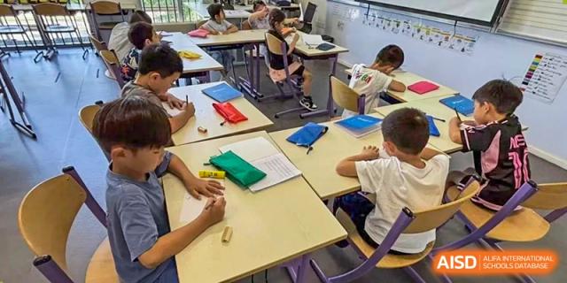 Students are actively working on assignments and taking notes while seated at yellow desks in a bright classroom at the Proviseur, du Lycée Français International de Pékin.