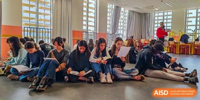 A group of older students from the Proviseur, du Lycée Français International de Pékin, are sitting on the floor in a brightly lit room, focused on reading and reviewing papers together.