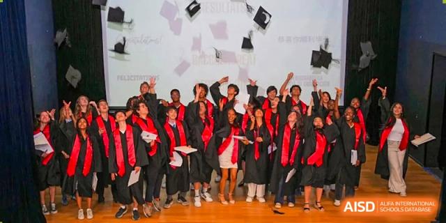 A joyful group of graduating students from the Proviseur, du Lycée Français International de Pékin, in red and black gowns, toss their caps high into the air during a ceremony.