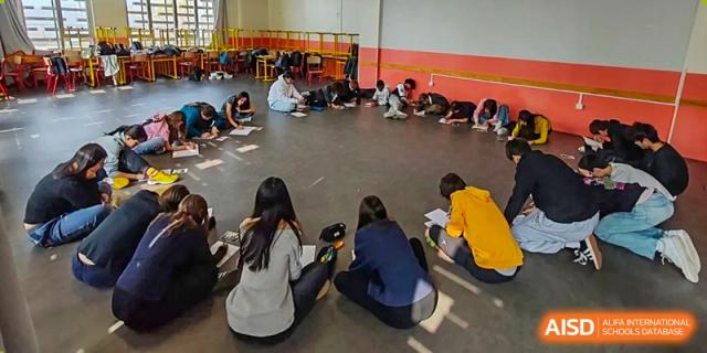 A small circle of students at the Proviseur, du Lycée Français International de Pékin, sits on the floor for a relaxed group activity or discussion session in a gym-like space.