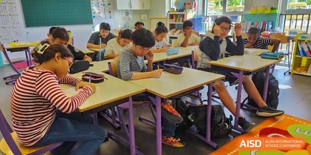 Students at the Proviseur, du Lycée Français International de Pékin, are busy writing and learning, seated at their desks in a classroom filled with natural light.