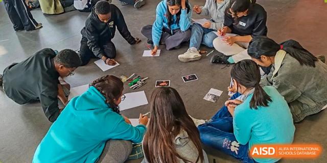 A small group of students from the Proviseur, du Lycée Français International de Pékin, sits on the floor, looking down at a paper as they collaborate on a task.