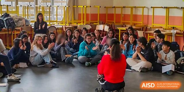 A large group of students from the Proviseur, du Lycée Français International de Pékin, is seated on the floor in a spacious, sunny indoor area for an assembly or presentation.