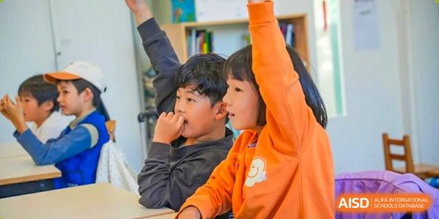 Two young students raising their hands to answer a question in a classroom setting at Proviseur, du Lycée Français International de Pékin.