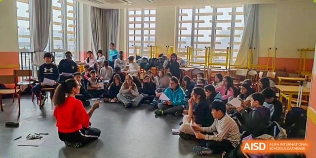 A wide shot shows a large number of students from the Proviseur, du Lycée Français International de Pékin, seated on the floor during a school-wide assembly or meeting.
