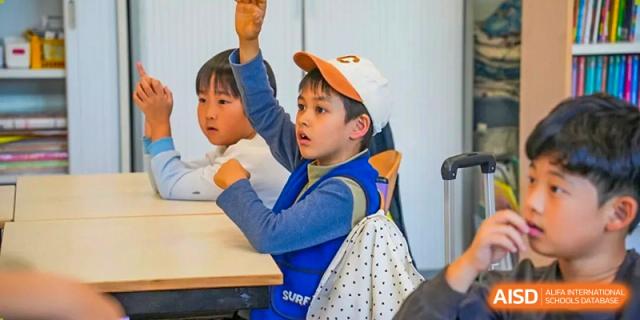 Boy in a white cap enthusiastically raising his hand in class next to a classmate at Proviseur, du Lycée Français International de Pékin.