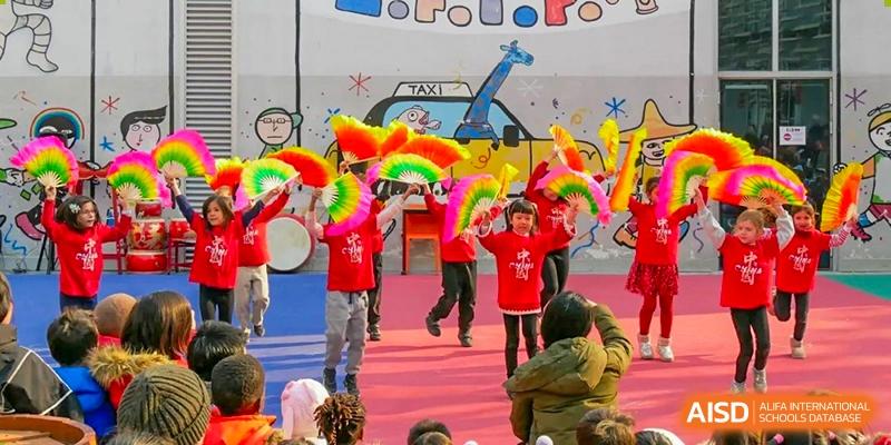 Children in vibrant, colorful costumes performing a traditional dance on an outdoor stage in front of a painted backdrop. Proviseur, du Lycée Français International de Pékin.