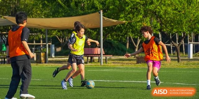 Two young girls in yellow vests and dark shorts actively playing soccer, maneuvering the ball across a sunny, grassy field. Proviseur, du Lycée Français International de Pékin.
