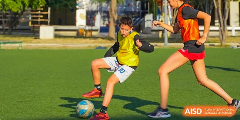 A young boy wearing a yellow vest and white shorts controls a soccer ball with his foot during an outdoor game on a green field. Proviseur, du Lycée Français International de Pékin.