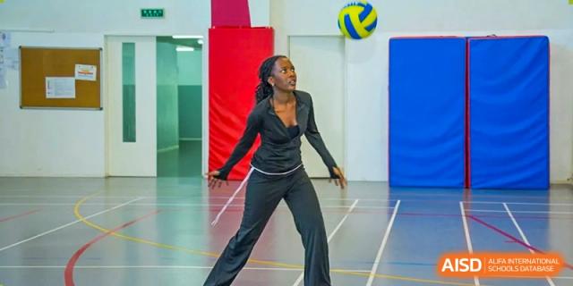 A young woman in a black outfit performing a dynamic dance move on a polished indoor sports court with a blue wall backdrop. Proviseur, du Lycée Français International de Pékin.