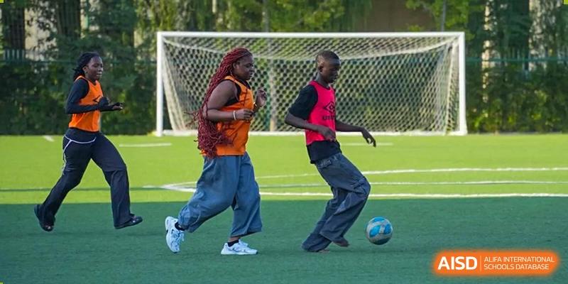 Two boys, one in an orange vest and the other in a black and red shirt, dribbling a soccer ball toward a goal on a turf field. Proviseur, du Lycée Français International de Pékin.