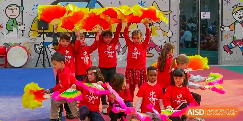 A group of children in red shirts and pink accents perform a choreographed routine with excited, raised hands. Proviseur, du Lycée Français International de Pékin.