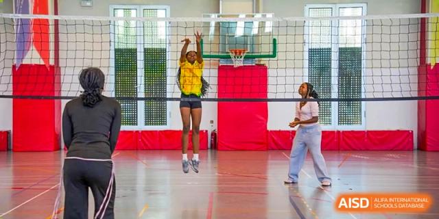 A girl in a yellow shirt jumps in the air on an indoor basketball court, with a teammate standing nearby. Proviseur, du Lycée Français International de Pékin.