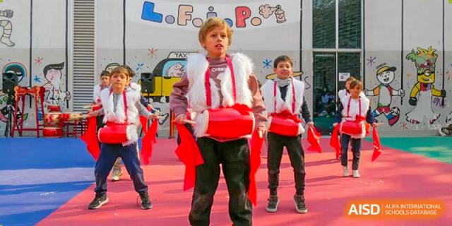 A boy in a white and red costume energetically drumming while participating in an outdoor performance with other children. Proviseur, du Lycée Français International de Pékin.