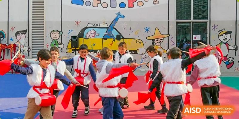 A group of children in white and red costumes drumming outdoors in front of a colorful, painted mural backdrop. Proviseur, du Lycée Français International de Pékin.