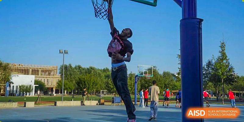 A person leaps high into the air to dunk a basketball on an outdoor court with a bright blue sky background. Proviseur, du Lycée Français International de Pékin.
