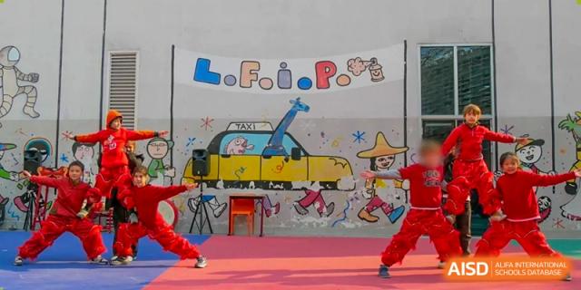 A group of children in red costumes performing a synchronized routine in front of a colorful backdrop featuring a yellow taxi. Proviseur, du Lycée Français International de Pékin.