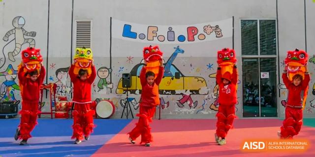 A child in red performing a traditional dance next to a drum, with a banner displaying 'L.F.I.P.' and a yellow taxi image in the background. Proviseur, du Lycée Français International de Pékin.