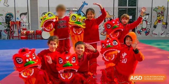 Children in red holding a lion head and smiling while performing a cultural dance on an outdoor stage. Proviseur, du Lycée Français International de Pékin.