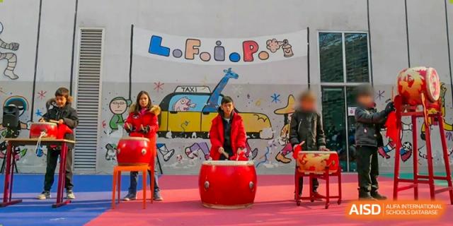 Two people next to a large red drum, with one person actively drumming during a cultural event under a banner. Proviseur, du Lycée Français International de Pékin.