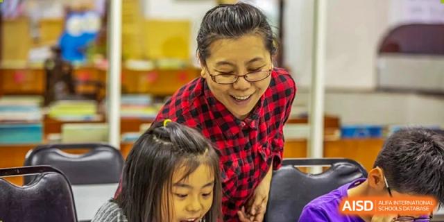 A teacher in a red plaid shirt smiles while working closely with a young student on a project at Tianjin International School (TIS).