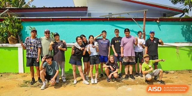 A group of students poses together outdoors in front of a colorful turquoise wall at Tianjin International School (TIS).