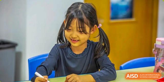 A young girl with pigtails smiling warmly while focusing on an activity during a classroom lesson at Tianjin International School (TIS).