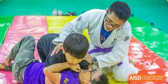 An instructor in a white gi guiding young students during a martial arts or grappling practice on a yellow mat at Tianjin International School (TIS).