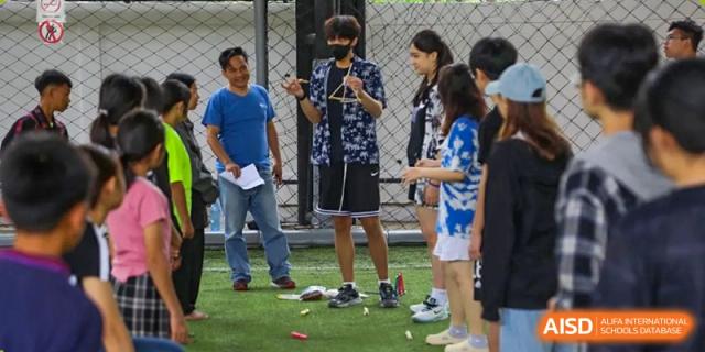 Students standing on an outdoor green field participate in a group activity or lesson with an instructor at Tianjin International School (TIS).