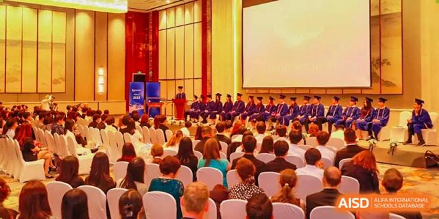 A large audience watches a formal graduation ceremony with students in purple robes on a brightly lit stage at Tianjin International School (TIS).