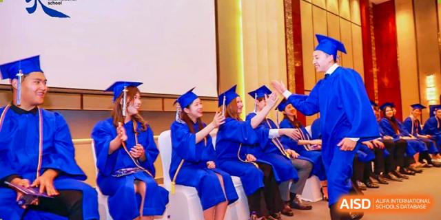 Graduating students in blue caps and gowns sitting in a row and high-fiving during a commencement ceremony at Tianjin International School (TIS).