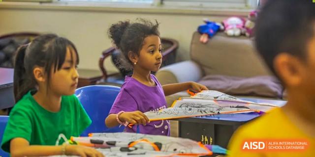 A young girl in a purple shirt proudly displaying her artwork or a colorful paper craft during an art lesson at Tianjin International School (TIS).