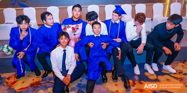 Three male graduates in blue gowns and caps sitting and laughing together during a formal school ceremony at Tianjin International School (TIS).