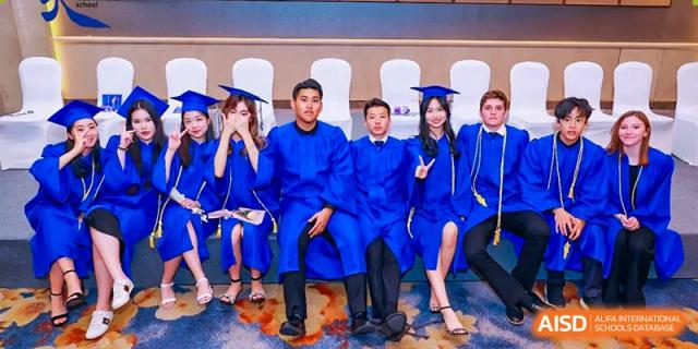 Four graduates in blue caps and gowns sitting in a row on stage, smiling and posing for a group photo at Tianjin International School (TIS).