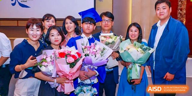 A smiling graduate in a blue cap and gown standing with his family and holding large bouquets of flowers at Tianjin International School (TIS).