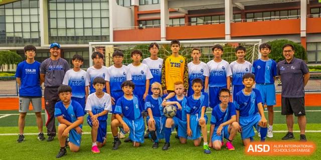 A boys' soccer team in blue and white jerseys poses for a group photo on a green field with their coaches at Tianjin International School (TIS).