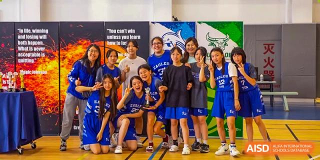 A group of girls in blue basketball uniforms cheer and pose together indoors, with one student holding a silver trophy for Tianjin International School (TIS).