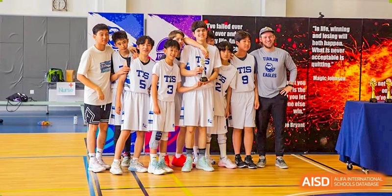 Members of a boys' basketball team in white "TIS" uniforms stand together on a court while holding a championship trophy at Tianjin International School (TIS).
