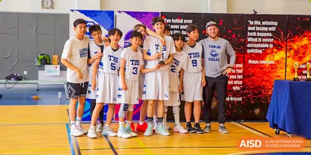 Members of a boys' basketball team in white "TIS" uniforms stand together on a court while holding a championship trophy at Tianjin International School (TIS).