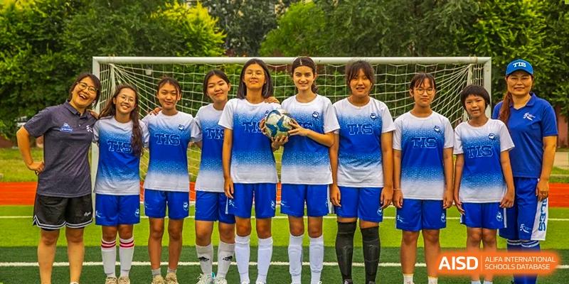 Four girls in blue soccer kits stand together on a grass field, with the center player holding a soccer ball at Tianjin International School (TIS).