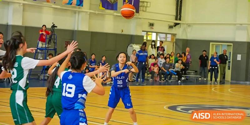 A girl in a blue basketball uniform prepares to receive a pass during a game in a crowded gymnasium at Tianjin International School (TIS).