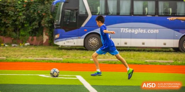 A young boy in a blue kit runs across a green soccer field with a team bus visible in the background at Tianjin International School (TIS).