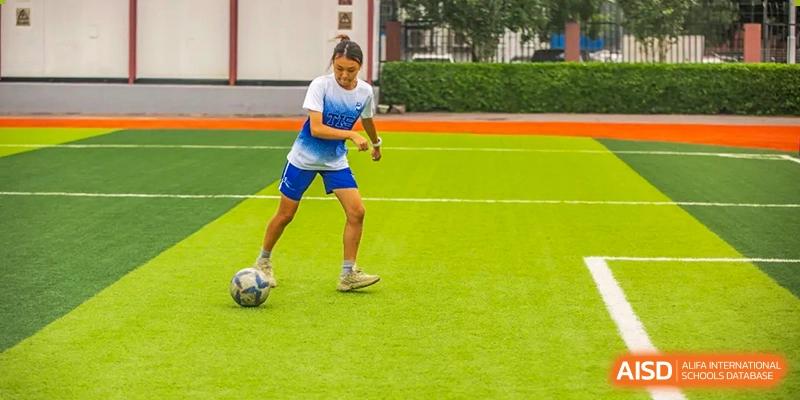 A girl in a white and blue soccer jersey dribbles a ball across a bright green turf field during practice at Tianjin International School (TIS).