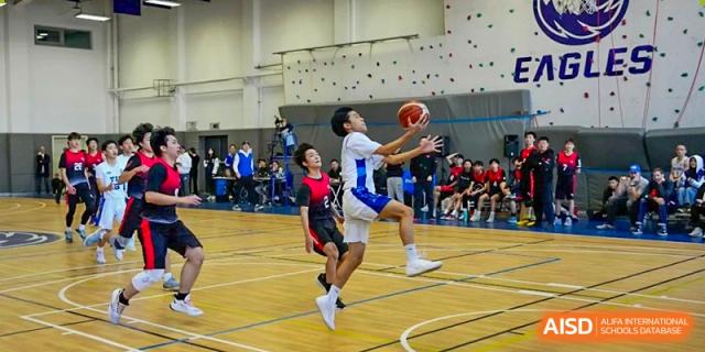 A student athlete jumps for a layup during a competitive basketball game in a gymnasium featuring a climbing wall at Tianjin International School (TIS).