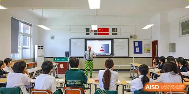 A teacher stands near a large screen, instructing students seated in a brightly lit classroom with a Chinese national flag displayed prominently on the wall at Maple Leaf International School Shanghai.