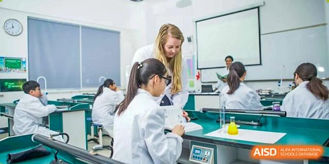 A female student in a lab coat performs an experiment at a desk, receiving helpful guidance from a female instructor in a busy science laboratory at Maple Leaf International School Shanghai.
