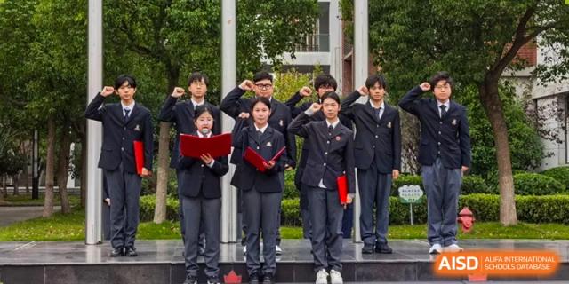 A small group of students in uniforms stands outdoors next to a flagpole, holding red folders and saluting during a formal ceremony at Maple Leaf International School Shanghai.