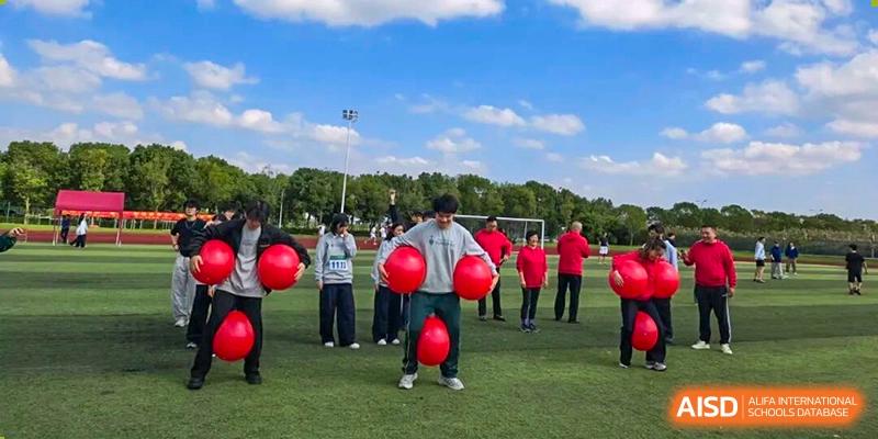 A large group of students, mostly dressed in red, stands on a sunny grass field holding red balloons for a cooperative team-building event at Maple Leaf International School Shanghai.