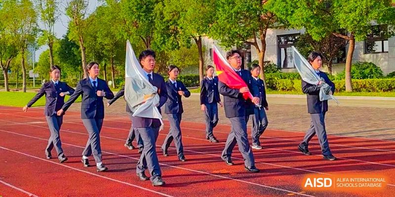 A group of students runs with enthusiasm on a red track, carrying flags as they participate in a school sports day parade or event at Maple Leaf International School Shanghai.