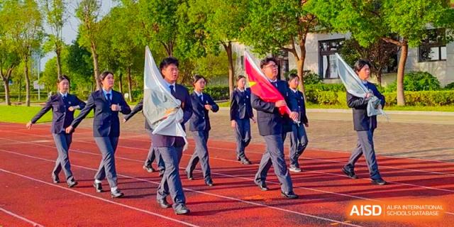 A group of students runs with enthusiasm on a red track, carrying flags as they participate in a school sports day parade or event at Maple Leaf International School Shanghai.